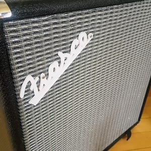 May include: A black guitar amplifier cabinet featuring a textured black exterior, a silver woven grille cloth, and a black top handle with chrome hardware. A white script logo reading "Fiasco" is mounted diagonally across the front grille.