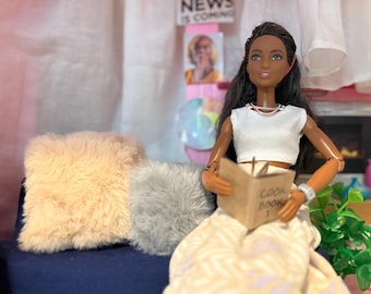 May include: A doll with long black hair wearing a white crop top and a patterned skirt, holding a small book labeled "Cook Book 1." Two fluffy pillows in beige and grey sit on a blue surface. A sign that says "NEWS IS COMING" is in the background.