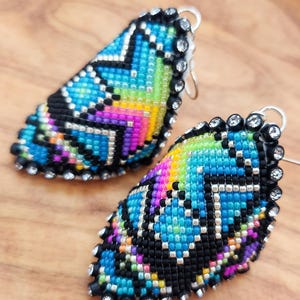 Op de afbeelding: Een paar kleurrijke kralen oorbellen met een geometrisch patroon en zilveren accenten. De oorbellen zijn traanvormig en hebben een zwarte achtergrond met blauwe, groene, roze en gele kralen.