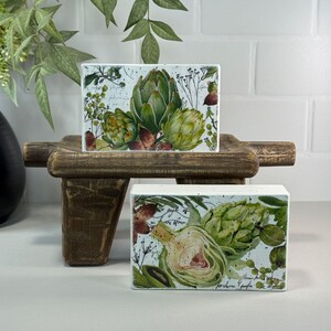 May include: Two rectangular decorative blocks with artichoke illustrations. The blocks feature green artichokes with watercolor-style details, set against a light blue background with botanical accents. The blocks are displayed on a rustic wooden stand.