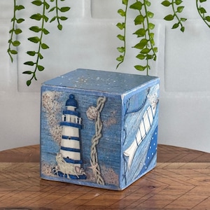 May include: A blue wooden box with a nautical theme. The box features a lighthouse, a rope, a seashell, and a fish. The box is painted with a blue and white striped pattern.