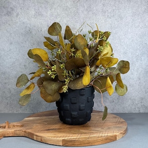 May include: A matte black ceramic pot with a textured grid pattern containing a dense arrangement of artificial eucalyptus branches. The foliage features rounded leaves in shades of olive green, mustard yellow, and brown with small green buds.