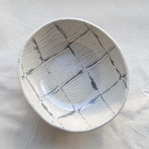May include: A white ceramic bowl with a gray, grid-like pattern. The bowl's interior features a series of thin, parallel lines, creating a textured effect. The bowl is empty and sits on a white surface.