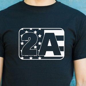 Pode incluir: Uma camiseta preta com um gráfico branco das letras "2A" dentro de um retângulo. O retângulo é preenchido com um design de bandeira americana estilizado.