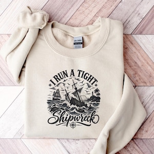 Könnte beinhalten: Ein cremefarbenes Sweatshirt mit Rundhalsausschnitt und einem schwarzen Grafikdruck eines Schiffswracks auf dem Meer. Das Design zeigt ein Segelschiff, einen Leuchtturm, Palmen und den Text I RUN A TIGHT SHIPWRECK mit einem Kompassrosensymbol.