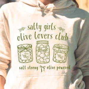 May include: Cream-colored hoodie with a green graphic. The graphic features three jars of olives and the text "salty girls olive lovers club". Below the jars are the words "salt strong" and "olive powered".