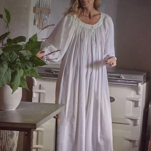 May include: A long, white nightgown with a round neckline and long sleeves. The nightgown has a button-down front and is made of a lightweight fabric. The woman is standing in a kitchen with a white stove and a potted plant.
