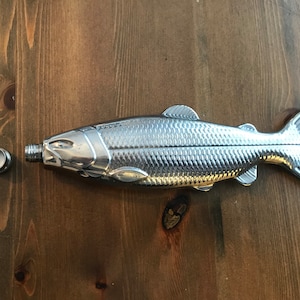 Puede incluir: Una petaca de plata con forma de pez con un tapón extraíble. La petaca está hecha de metal y tiene un diseño detallado.