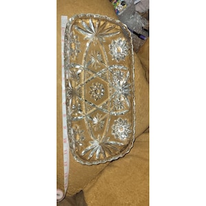 May include: A clear, rectangular pressed glass tray featuring a geometric starburst pattern. A white measuring tape is positioned along the side, indicating the tray has a length of approximately 14 inches. The glass has a scalloped edge.