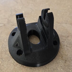 May include: A black 3D-printed object with a circular base and two vertical supports. The supports have intricate, curved designs. The base has a central hole and several smaller holes around the perimeter. The object is made of black plastic.