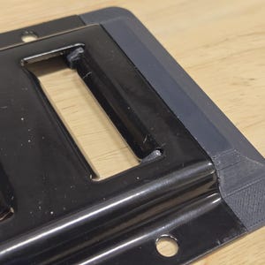 May include: A black metal plate with a rectangular cutout and a gray plastic edge. The plate has two small holes on the bottom.