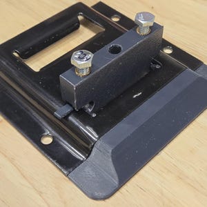 May include: A black metal bracket with a rectangular base and a raised section, secured with two silver bolts. The bracket has a dark gray plastic edge and is designed for mounting or support.