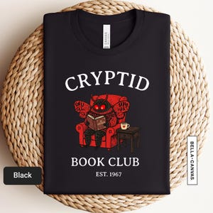 Op de afbeelding: Zwart T-shirt met de witte tekst "CRYPTID BOOK CLUB EST. 1967". De afbeelding toont een roodoogmotwezen dat een boek leest in een rode fauteuil, met een tafeltje en een kopje koffie. Het shirt is gemaakt van Canvas materiaal.