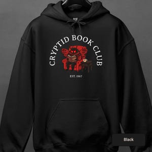 May include: Black hooded sweatshirt with a graphic of a red monster reading a book in a red armchair. The text "CRYPTID BOOK CLUB" arches above the illustration, with "EST. 1967" below. A coffee cup sits on a small table. The hoodie has a drawstring hood.