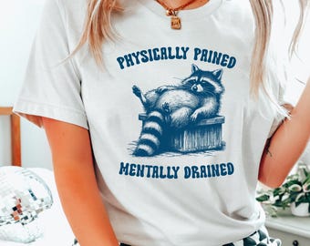 Puede incluir: Camiseta blanca con un gráfico azul de un mapache sentado en una caja. El texto "PHYSICALLY PAINED" y "MENTALLY DRAINED" está impreso en la camiseta.