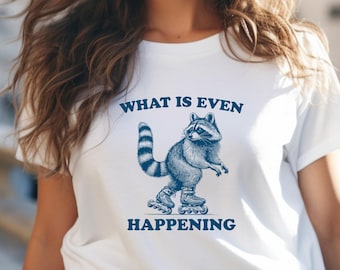 May include: White t-shirt with a blue graphic of a raccoon wearing roller skates. The text "What is even happening" is printed on the shirt.