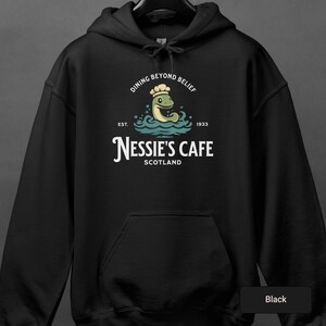 Puede incluir: Sudadera con capucha negra con un gráfico blanco de un Nessie de dibujos animados con un gorro de cocinero y sosteniendo una espátula. El gráfico dice "Dining Beyond Belief" y "Nessie's Cafe Scotland" con las fechas "Est. 1933".