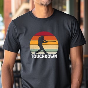 Puede incluir: Una camiseta gris oscuro con un gráfico de estilo vintage de un jugador de béisbol golpeando una pelota. El gráfico está sobre un fondo de puesta de sol con el texto "TOUCHDOWN" debajo.