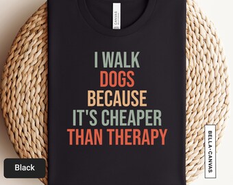 以下が含まれることがあります： レトロなフォントで「I walk dogs because it's cheaper than therapy」と書かれた黒いTシャツです。