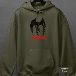 May include: Military green hoodie with a black graphic of a winged creature with red eyes and the word "MOTHMAN" in red. The hoodie has a drawstring hood and a front pocket.