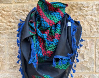 May include: Black scarf with a colorful geometric pattern in red, green, and blue. The scarf has blue tassels along the edges. The scarf is draped over a black form, showcasing its design and texture.