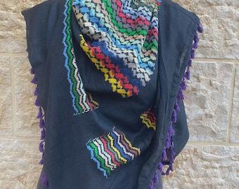 May include: A black scarf with a colorful, patterned design and purple tassels. The scarf features a geometric pattern in shades of red, green, blue, and yellow. The edges are trimmed with purple tassels.