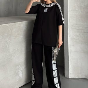 Op de afbeelding: Zwart tweedelig outfit met een top met korte mouwen en een broek met wijde pijpen. De top en broek hebben witte patroonafwerking. De outfit wordt gedragen met witte sneakers en een zonnebril. De persoon houdt een telefoon en een krant vast.