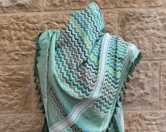 May include: A light green scarf with a zigzag pattern in shades of green, blue, and red. The scarf has white stripes and green tassels along the edges. The scarf is draped over a person.