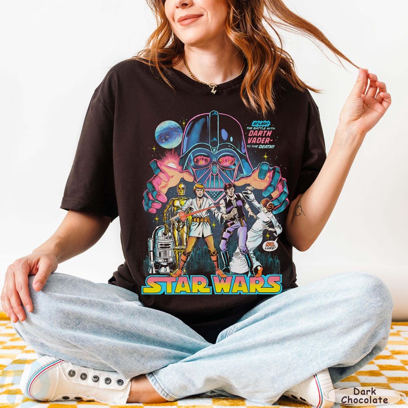 May include: Dark chocolate-colored Star Wars graphic tee featuring Darth Vader and other characters. The design includes the text "STAR WARS" in yellow and comic book-style illustrations. The shirt is a short-sleeved, crew neck style.