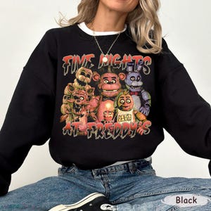 May include: Black crewneck sweatshirt featuring a graphic from the video game Five Nights at Freddy's. The design includes several animatronic characters and the game's title in a distressed font. The sweatshirt has a white collar.