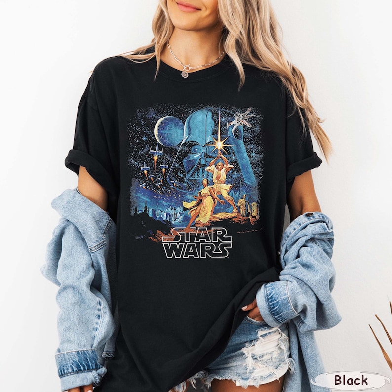 Pu&ograve; includere: T-shirt nero con un design di poster del film Star Wars. La grafica include personaggi, astronavi e il logo di Star Wars in bianco. Il design &egrave; ambientato su uno sfondo blu e stellato.