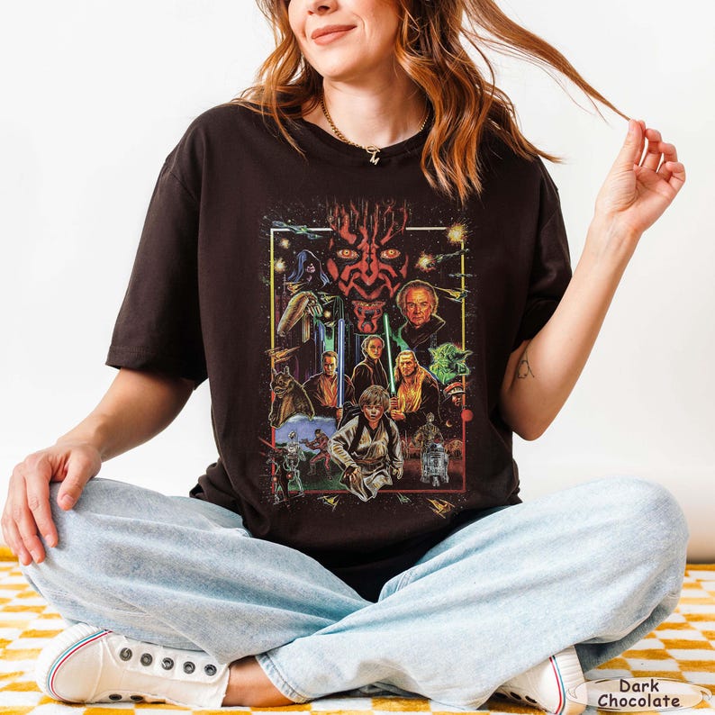 May include: Dark chocolate-colored t-shirt featuring a Star Wars graphic. The graphic includes various characters and scenes from the Star Wars saga, with a prominent image of Darth Maul. The shirt is a crew neck style.