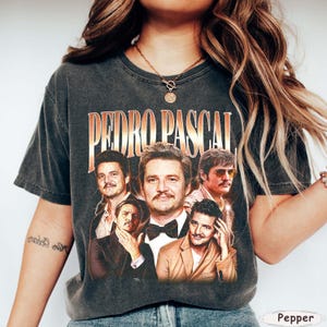 May include: Dark gray t-shirt featuring a collage of images of Pedro Pascal with the text "PEDRO PASCAL" in a gold and orange font. The shirt has a vintage, worn look.