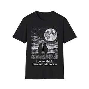 May include: A black short-sleeve t-shirt featuring a white graphic print of two cats sitting on a stone wall under a full moon and starry night sky. Below the illustration, the text reads: i do not think therefore i do not am.