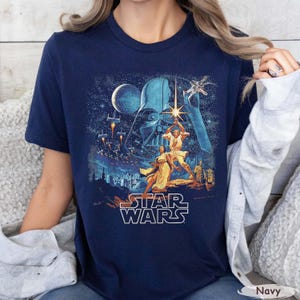 Pu&ograve; includere: T-shirt blu navy con grafica di Star Wars. Il design include Darth Vader, Luke Skywalker, la Principessa Leia e varie astronavi. Le parole "STAR WARS" sono visualizzate in un carattere bianco stilizzato.