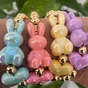 Puede incluir: Un conjunto de cinco pulseras de colores pastel con dijes de conejito y cuentas en forma de corazón. Las pulseras están hechas de cuentas doradas y presentan una variedad de colores, incluyendo azul claro, rosa, amarillo y morado.