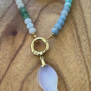 Puede incluir: Un collar de cuentas con un cierre dorado y un colgante. El collar presenta un degradado de cuentas azules, verdes y blancas. El colgante es un dije en forma de pez, de color lila claro. El collar se exhibe sobre una superficie de madera.