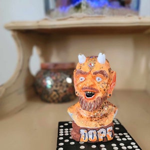 Puede incluir: Figurita naranja y dorada de un demonio con barba y cuernos. La figurita está sentada en un tablero de dominó blanco y negro con la palabra "DOPE" escrita en él.