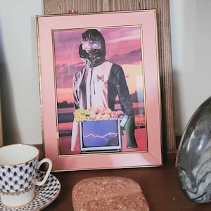 Puede incluir: Una impresión enmarcada de una persona que lleva una chaqueta negra y blanca con un diseño de mariposa. La persona está de pie frente a un televisor con un rayo en la pantalla. El fondo es una puesta de sol rosa y púrpura.