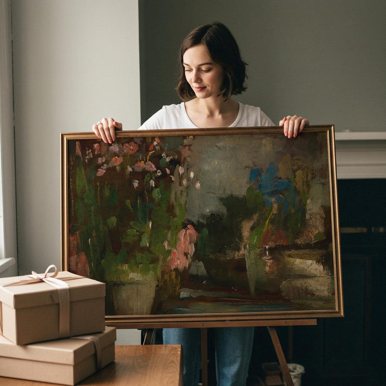 May include: A rectangular oil painting in a thin gold frame featuring an impressionistic landscape with dark green foliage, soft pink floral accents, and a muted blue sky. The artwork is displayed on a wooden easel, showcasing visible brushstrokes.