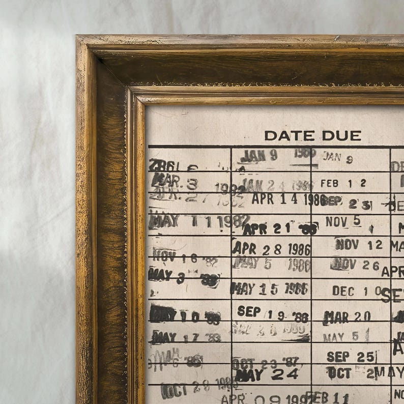 May include: A framed vintage library card pocket with a gold-colored wooden frame. The card pocket has the words "DATE DUE" at the top and contains handwritten dates and numbers. The frame has a distressed, antique look.