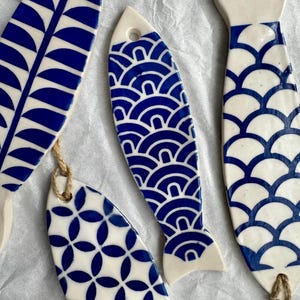 May include: Several ceramic fish ornaments with blue and white patterns. Each fish has a unique design, including geometric shapes and wave-like patterns. They are strung with twine for hanging.