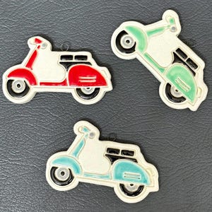 May include: Three ceramic scooter ornaments in red, green, and blue. Each scooter has a white body with black accents and a small loop for hanging. The scooters are on a dark gray surface.