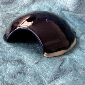 May include: A dark blue ceramic reptile hide with a rounded opening. The hide is glazed and has a smooth, shiny finish.