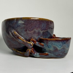 May include: A ceramic bowl with a smaller attached side compartment, featuring a glossy glaze in shades of deep purple, blue, and brown. The surface has a mottled, blended appearance with smooth, rounded edges and a continuous, flowing form.