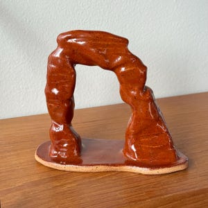 May include: A ceramic sculpture of a natural arch, glazed in a reddish-brown color. The arch is supported by two pillars and sits on a rectangular base. The sculpture is displayed on a wooden surface.