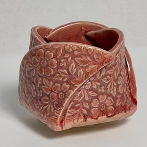 May include: A small, hand-formed ceramic bowl with a dusty rose glaze. The exterior features a deeply embossed, intricate floral pattern of flowers and leaves. The rim is folded and irregular, creating a sculptural, organic shape with a glossy finish.