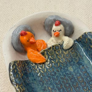 May include: A ceramic dish featuring two sleeping birds under a blue blanket. One bird is orange, the other is white, both with red accents. The dish has a white rim and a textured blue interior.