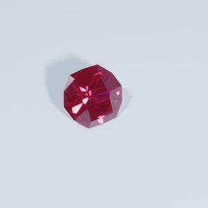 May include: A faceted, deep red gemstone with a geometric shape. The gem is set against a white background, with a shadow cast to the left. The gem's facets reflect light, creating a sparkling effect. This could be a ruby or similar precious stone.