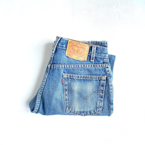 May include: Blue denim jeans with a classic five-pocket design. The jeans feature a faded wash and a leather patch on the back waistband. The back pocket has a small red tag. The jeans are folded neatly on a white background.